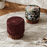 Two cylindrical upholstered ottomans on a patterned rug in a styled interior. The front ottoman has a dark red textured fabric top and a fluted brown wooden base.