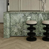 Black pedestal bar stools with cream upholstered seats in front of a green veined stone kitchen island.