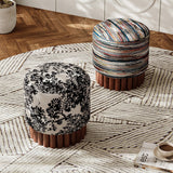 Two round upholstered ottomans with fluted brown wooden bases on a patterned rug in a styled interior. One ottoman has a black floral toile-style print on a light beige background, and the other has multicolored horizontal striped upholstery.
