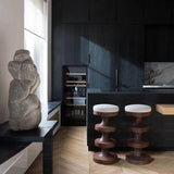 Two brown wooden bar stools with cream cushions beside a black kitchen island near a window and stone sculpture.