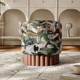 A cylindrical upholstered ottoman with a detailed landscape pattern in green, beige, and brown, resting on a fluted reddish-brown wooden base in a styled room setting.