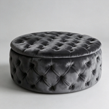 A round, tufted velvet ottoman stool in dark gray, shown from a three-quarter angle against a neutral light gray background.