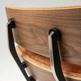 Close-up of a modern wooden chair showing the backrest, upholstered seat, and black metal frame details against a light gray background.