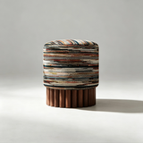 A cylindrical upholstered stool with a multicolored patterned fabric top and a fluted dark wood base, photographed on a light gray studio background.