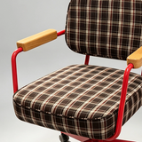 A close-up of an office chair with a brown, beige, and white plaid upholstered seat and backrest, supported by a red metal frame with light wood armrests.