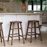 Brown wooden bar stools with round seats and turned legs beside a white kitchen island with brass taps.
