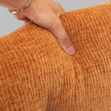 Close-up of a person's hand pressing down on the textured orange-brown corduroy fabric of a cushion or pillow, showing the fabric's ribbed weave and color.