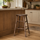 Brown wooden bar stool with a round seat and turned legs beside an oak-toned kitchen island with a pale stone top.