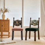 Two dark wood chairs with pink and green seats and patterned backrests beside a cabinet in a sunlit room.