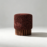 A cylindrical upholstered stool or ottoman with a textured burgundy and rust fabric top and a fluted wooden pedestal base, photographed on a light gray studio background.
