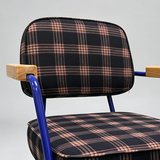 A close-up of a chair with a black and pink plaid upholstered seat and backrest, blue metal legs, and light wood armrests against a light gray background.