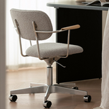 Rear three-quarter view of a taupe boucle office chair beside a black desk, showing its metal frame and castor base.