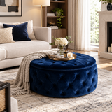 A round tufted ottoman in deep navy blue velvet, styled in a bright, modern living room with a beige sofa, a throw blanket, and a decorative tray on top.