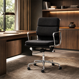 A modern black fabric office chair with a chrome five-star base and casters, positioned in a stylish, contemporary home office setting with a wooden desk, lamp, and warm wood shelving in the background.