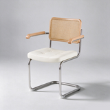 A modern cantilever dining chair with a white tufted seat cushion, a natural woven cane backrest, light wood armrests, and a polished chrome metal frame, shown against a neutral gray studio background.