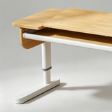 A close-up studio shot of a modern desk with a light natural wood top and a white metal pedestal base, showing the underside support and leg adjustment area.