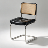 A modern dining chair with a black tufted leather seat, a natural woven cane backrest, and a chrome cantilever steel base, shown on a light gray studio background.