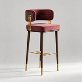 A modern bar stool with a deep burgundy velvet upholstered seat and curved backrest, dark brown wood legs and frame, and polished gold metal accents on the legs and footrest, set against a neutral light gray background.