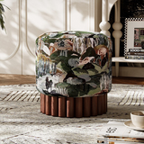 A round upholstered ottoman stool with a multicolor botanical tapestry pattern on the cushion and a fluted dark wood base, placed on a patterned rug in a styled interior room.