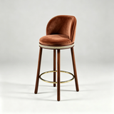 Brown Alma bar stool with a curved upholstered backrest, dark wood legs and a gold-tone circular footrest.
