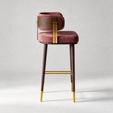 A modern bar stool featuring a dusty rose velvet cushioned seat and backrest, dark wood legs and backrest trim, and polished gold metal accents on the frame and leg caps, set against a neutral light gray background.