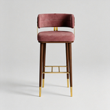 A modern bar stool featuring a dusty rose velvet upholstered seat and backrest, dark brown wooden legs, and polished gold metal accents including the armrests, footrest, and leg caps, set against a neutral light gray background.