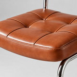 Close-up of a tufted brown leather stool seat with a polished chrome metal leg visible against a light gray background.