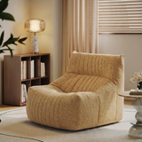 A plush, ribbed fabric armchair in a warm beige/tan color, styled in a cozy, modern living room setting next to a wooden bookshelf and a lit table lamp.