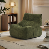 A plush, low-profile lounge chair upholstered in textured olive green bouclé fabric, styled in a warm, modern living room setting with a wooden bookshelf, table lamp, and neutral rug.
