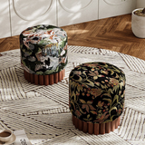 Two round upholstered ottomans with fluted brown wooden bases are placed on a patterned rug in a sunlit room. The left ottoman has a multicolor landscape print on a dark background, and the right ottoman has a black floral textile with gold, green, and red botanical motifs.