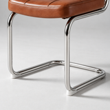 Close-up of a cantilever chair with a brown leather seat cushion and a polished chrome metal frame on a light gray studio background.