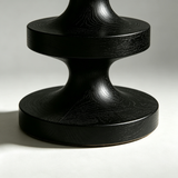 Close-up of the black stool's sculpted pedestal showing curved sections, circular tiers and visible wood grain.