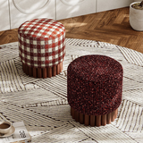 Two cylindrical upholstered ottomans with fluted wooden legs on a patterned rug in a styled interior. One ottoman has a red and white gingham pattern, and the other has a dark red and black speckled texture.
