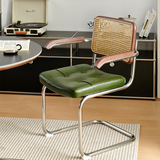 A modern cantilever dining chair with a green tufted leather seat, a woven cane backrest, and rose gold metal armrests, set in a styled room with a wooden floor, a black sideboard, and a rug.