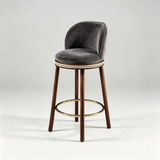 Charcoal grey Alma bar stool with a padded round seat, curved backrest, dark wood legs and gold-tone footrest.
