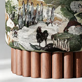 Close-up of a cylindrical upholstered stool or ottoman with a woven landscape pattern in muted green, beige, brown, and black, supported by a fluted base made of vertical wooden legs.