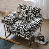 A modern armchair with a bold black and white zebra print upholstery and chrome metal legs, styled in a bright room with a window, floor lamp, and patterned rug.