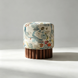 A cylindrical upholstered stool with a cream fabric top featuring a botanical pattern in blue, green, and orange, supported by a fluted dark brown wooden base, set against a neutral gray background.