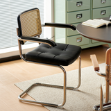 A modern cantilever dining chair with a black faux leather seat and backrest, a woven cane back panel, and a chrome metal frame, shown in a bright room next to a round table.