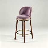Mauve Alma bar stool with a curved upholstered backrest, dark wood legs and a gold-tone circular footrest.
