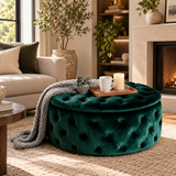 A round tufted velvet ottoman in deep emerald green sits in a styled living room. A gray knit throw blanket and a small wooden tray with two white mugs and a candle are placed on top.