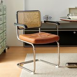 A modern cantilever dining chair with a brown leather seat, woven cane backrest, and chrome metal frame, set in a bright, contemporary home office environment.