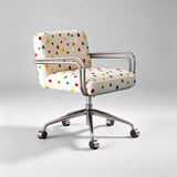 A modern armchair with a cream-colored fabric upholstered seat and backrest, decorated with a colorful polka dot pattern, supported by a polished chrome five-star base with casters, set against a neutral gray studio background.