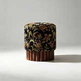 A cylindrical upholstered stool or ottoman with a black velvet fabric covered in an ornate floral pattern in gold, olive, and rust tones, set on a fluted dark wood base against a neutral light gray studio background.