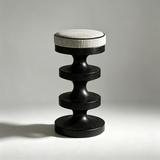 Black wooden bar stool with a cream upholstered round seat, black piping and a sculptural stacked pedestal base.