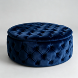 A round tufted velvet ottoman stool in deep navy blue, shown from a three-quarter angle against a neutral light gray background.
