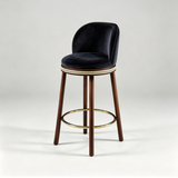 Black Alma bar stool with a padded round seat, curved backrest, dark wood legs and a gold-tone footrest.
