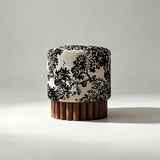 A cylindrical upholstered stool or ottoman with a black toile-style floral pattern on a warm beige fabric, set on a fluted dark brown wooden base against a light gray studio background.