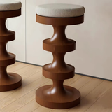 Brown wooden bar stool with a cream herringbone cushion and stacked curved pedestal, reflected in a nearby mirror.