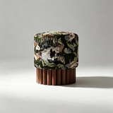 A cylindrical upholstered ottoman or stool with a multicolor botanical and animal print fabric on the upper half and a fluted medium-brown wooden base, photographed on a light gray studio background.
